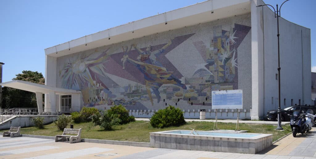 The façade of the Mangalia House of Culture featuring the monumental "Geneze" mosaic on a sunny summer day.